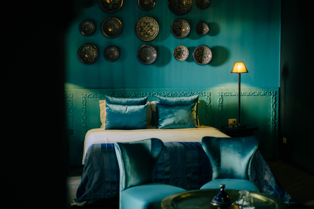 Close-up of a luxurious Moroccan bedroom detail, showing a carved wooden nightstand, elegant lamp, and rich textiles, moody lighting